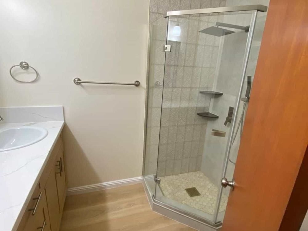 Puerta de ducha de esquina con paredes de vidrio, azulejos en tonos neutros, accesorios de baño modernos, en un baño funcional y bien diseñado por M&X Construction.